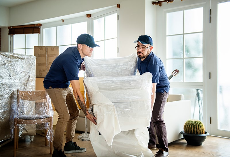 Two Workers In Uniform Carefully Move A Wrapped Piece Of Furniture In A Bright, Clean Living Room Setting, Showcasing San Antonio Property Cleanout And Eviction Cleaning Services. Two workers in uniform carefully move a wrapped piece of furniture in a bright, clean living room setting, showcasing San Antonio property cleanout and eviction cleaning services.
